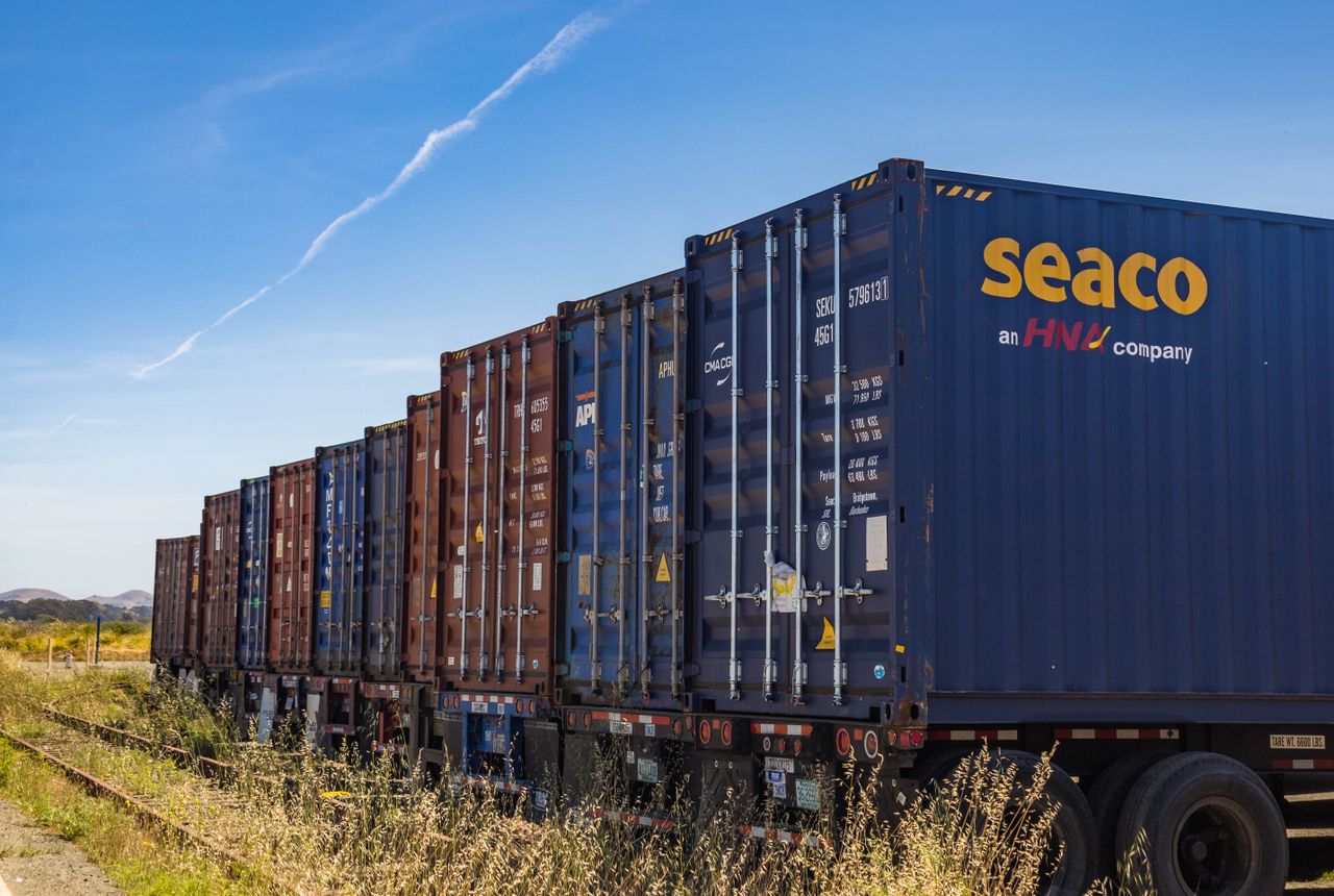 about-01 Row of vibrant cargo containers on train tracks under a clear blue sky.