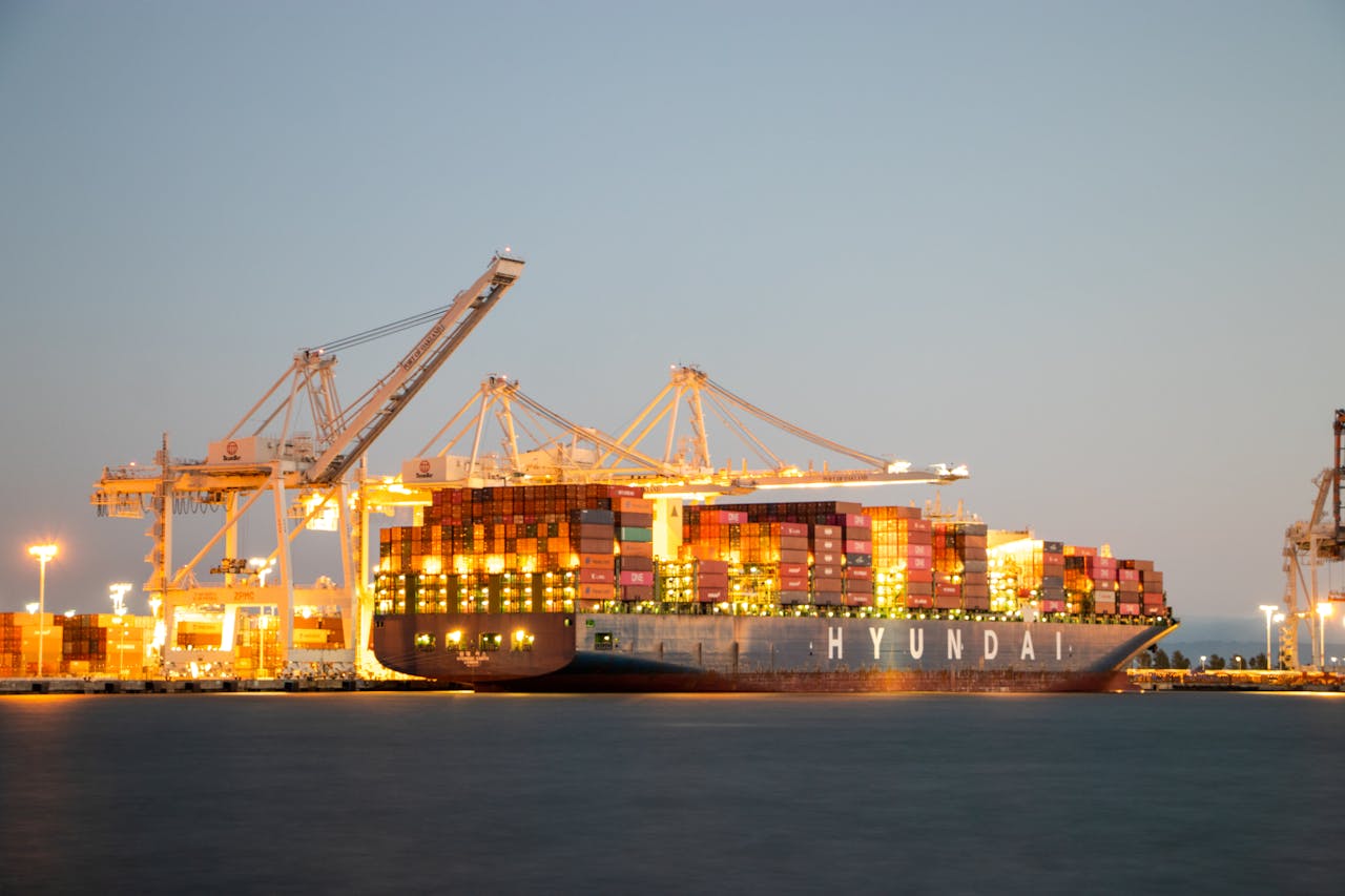 gallery-3 Container ship docked at port under cranes during twilight, illuminated against the evening sky.